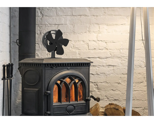 Een zwarte kachelventilator met vijf bladen op een brandende gietijzeren houtkachel voor een witte bakstenen muur met haardhout.