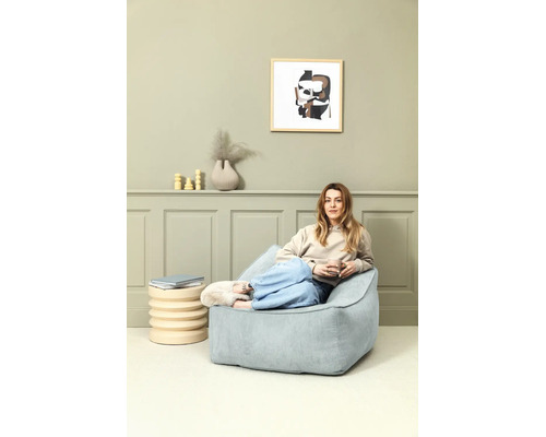 Vrouw zit ontspannen op een lichtblauwe corduroy zitzak in een modern ingerichte woonkamer met groene wandpanelen.