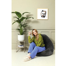Vrouw zit in een donkergrijze zitzak naast een grote kamerplant in een lichtgroen ingerichte kamer met wandpanelen.