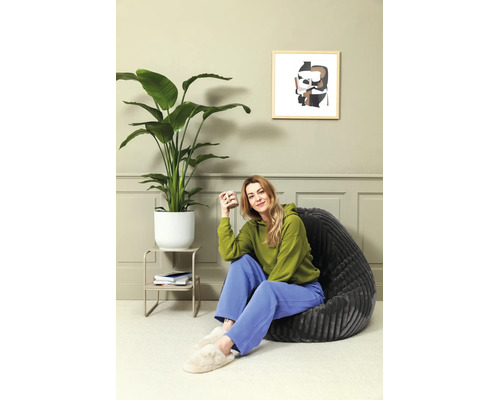 Vrouw zit in een donkergrijze zitzak naast een grote kamerplant in een lichtgroen ingerichte kamer met wandpanelen.