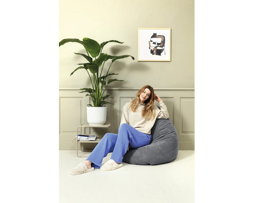 Een persoon zit in een grijze corduroy zitzak in een lichte woonkamer. Naast de zitzak staat een grote kamerplant in een witte pot.
