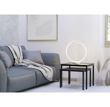 Moderne ringvormige LED-tafellamp op zwarte bijzettafels naast een grijze bank in een lichte, minimalistische woonkamer.