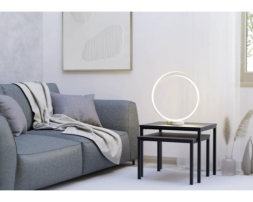 Moderne ringvormige LED-tafellamp op zwarte bijzettafels naast een grijze bank in een lichte, minimalistische woonkamer.