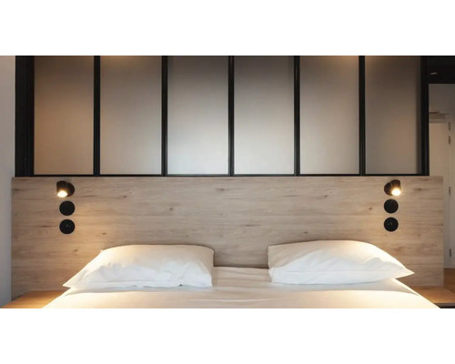 Moderne slaapkamer met hoofdbord in lichte houtlook, zwarte leeslampen, witte kussens en een glazen scheidingswand.