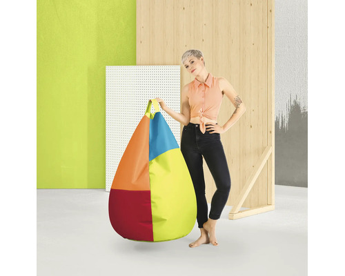 Een vrouw staat naast een grote, druppelvormige, veelkleurige zitzak met handvat in een moderne studio-omgeving met houten elementen.