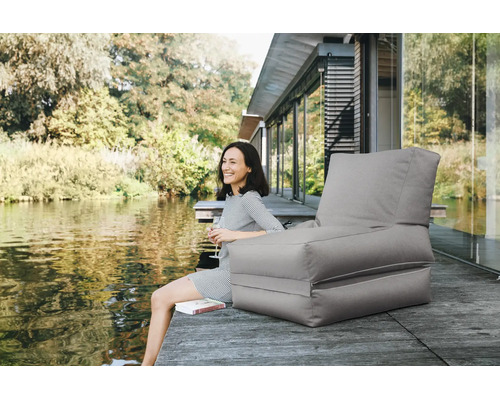 Grijze outdoor zitzak met rugleuning op een houten steiger aan het water, met daarnaast een glimlachende vrouw en een boek.