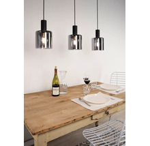 Drie hanglampen met cilindrische kappen van rookglas hangen boven een gedekte houten tafel in een lichte eetkamer.