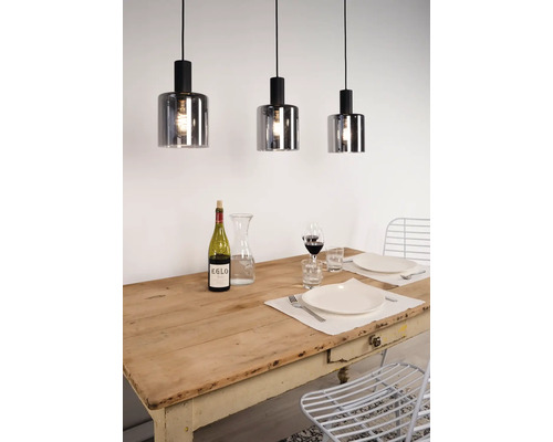 Drie hanglampen met cilindrische kappen van rookglas hangen boven een gedekte houten tafel in een lichte eetkamer.