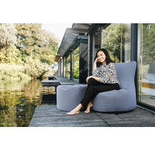 Een vrouw zit op een grijze outdoor zitzak met bijbehorende poef op een houten terras aan het water voor een modern huis.