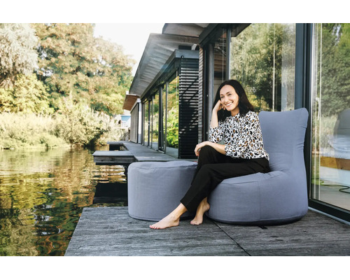 Een vrouw zit op een grijze outdoor zitzak met bijbehorende poef op een houten terras aan het water voor een modern huis.