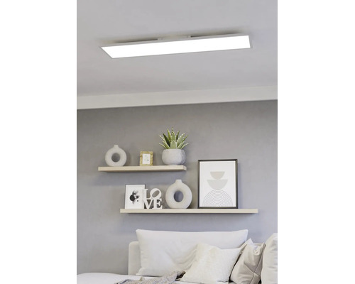 Modern, rechthoekig LED-paneel aan een wit plafond boven een lichte woonkamer met wandplanken en decoratieve elementen.