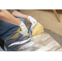 Een persoon met beschermende handschoenen snijdt minerale wol isolatie met aluminiumfolie met een isolatiemes.