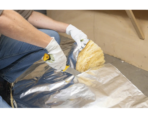 Een persoon met beschermende handschoenen snijdt minerale wol isolatie met aluminiumfolie met een isolatiemes.