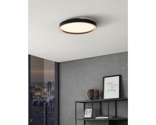 Platte, ronde zwarte LED-plafondlamp aan een wit plafond in een modern interieur met grijze wand en metalen rek.