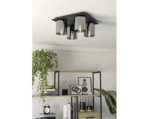 Vierlichts plafondlamp met zwarte metalen basis en cilindrische kappen van rookglas in een lichte woonkamer met een open kast.