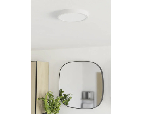 Ronde, platte LED-plafondlamp gemonteerd in een lichte kamer met spiegel en plant.