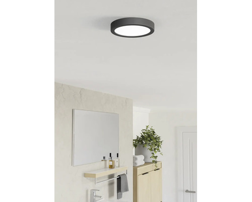 Ronde LED-plafondlamp met donker frame in een lichte, moderne badkamer met spiegel, groene plant en houten kast.