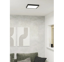 Vierkante LED plafondlamp met zwart frame aan een wit plafond in een moderne woonkamer met grijze muur en bank.