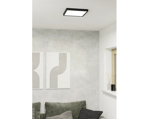 Vierkante LED plafondlamp met zwart frame aan een wit plafond in een moderne woonkamer met grijze muur en bank.