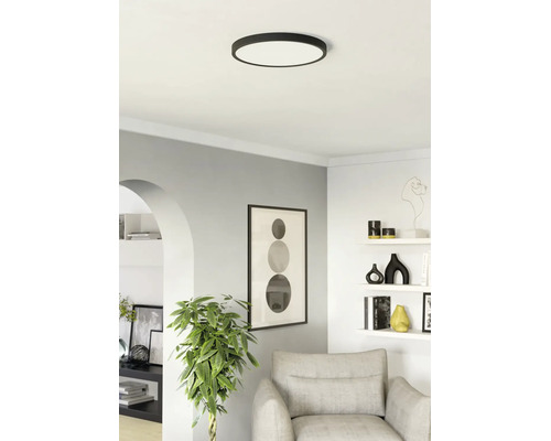 Platte, ronde plafondlamp in een moderne, lichte woonkamer met een grijze fauteuil, planken en een grote kamerplant.