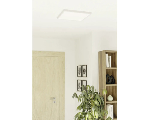 Vlak, vierkant ledpaneel aan het plafond in een lichte, modern ingerichte kamer met een houten deur, boekenkast en kamerplant.