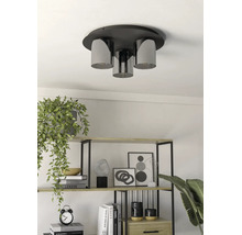 Drieliichts plafondlamp met cilindrische glazen kappen in een lichte, modern ingerichte kamer boven een plank met boeken en planten.