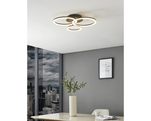 Moderne LED-plafondlamp met vier lichtgevende ringen in een lichte kamer met grijze muur, uitzicht op de stad en een witte tafel.