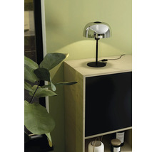 Moderne tafellamp met chromen kap en zwarte voet op een licht houten dressoir voor een lichtgroene muur.