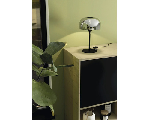 Moderne tafellamp met chromen kap en zwarte voet op een licht houten dressoir voor een lichtgroene muur.