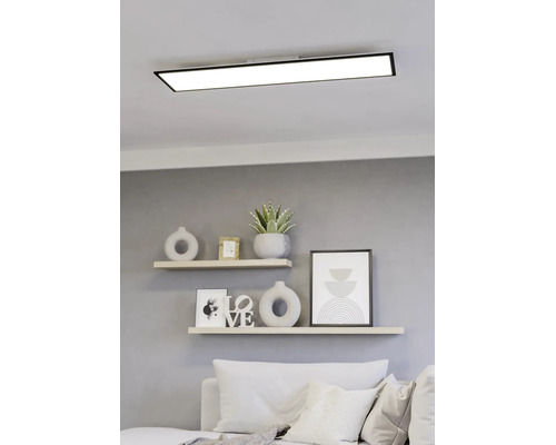 Rechthoekig LED-paneel met zwart frame aan het plafond boven een lichte, modern ingerichte woonruimte met planken en een bank.