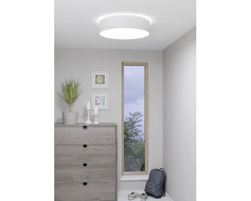 Ronde witte plafondlamp in een moderne kamer met een ladekast, een plant en een smal raam.