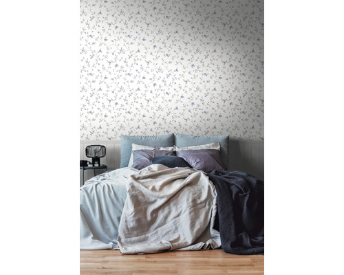 Lichte slaapkamer met bloemenbehang met fijne ranken en kleine bloemen. Het bed is opgemaakt met diverse kussens en dekens.