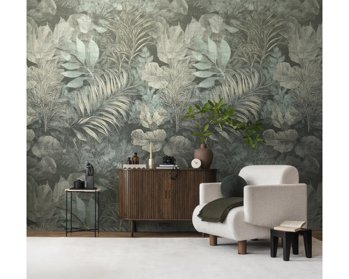 Vliesbehang met een groot, tropisch bladerpatroon in gedempte groentinten in een modern ingerichte woonkamer met dressoir en fauteuil.