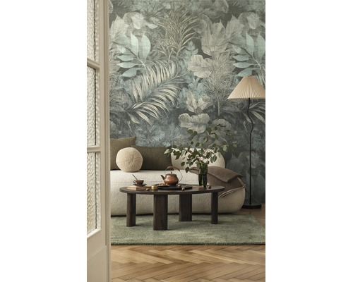 Vliesbehang met botanisch bladmotief in een woonkamer met een lichte bank, een donkere salontafel, een vloerlamp en een groen vloerkleed.