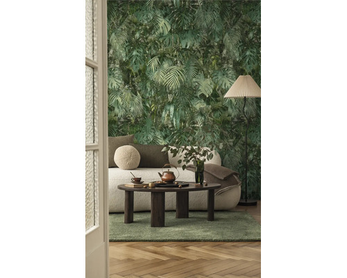 Vliesbehang met jungle-motief van palmbladeren en monstera in een woonkamer met een lichte bank, donkere houten tafel en een groen tapijt.