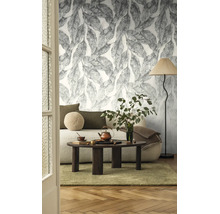 Woonkamer met vliesbehang met groot bladpatroon, bank met kussens, salontafel van donker hout en een vloerlamp.