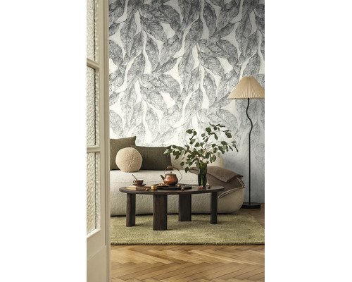 Woonkamer met vliesbehang met groot bladpatroon, bank met kussens, salontafel van donker hout en een vloerlamp.