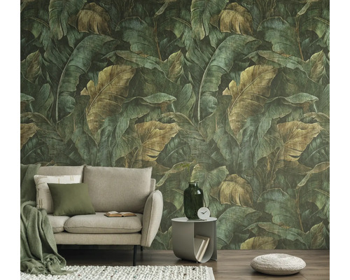 Vliesbehang met een groot junglepatroon van groene en bruine bladeren achter een beige bank in een woonkamer.