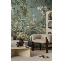 Fotobehang met botanisch motief van grote groene bladeren, kleurrijke vogels en bloemen in een licht ingerichte woonkamer met fauteuil.