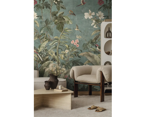 Fotobehang met botanisch motief van grote groene bladeren, kleurrijke vogels en bloemen in een licht ingerichte woonkamer met fauteuil.