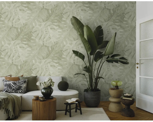 Lichte woonkamer met vliesbehang met monstera-bladmotief, bank, grote kamerplant en decoratieve houten bijtafels.