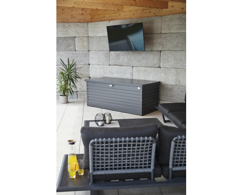 Metalen opbergbox in antraciet in een moderne loungehoek met stenen wand, televisie en tuinmeubels.