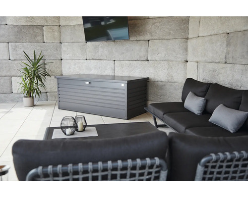 Metalen kussenkast op een licht terras met loungebank, bijzettafel, een plant en een muur van steenblokken.