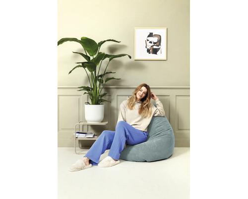 Een vrouw zit op een blauwe zitzak in een lichte, moderne kamer met een grote kamerplant en een abstract schilderij.