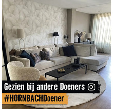 Modern ingerichte woonkamer met een lichte hoekbank, fauteuil, behang met bladmotief, rond vloerkleed, zwarte salontafel en Instagram Logo.