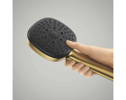 Een hand houdt een vierkante handdouche in goudkleur vast met een zwarte sproeikop en antikalknoppen.