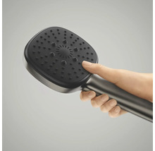 Een hand houdt een moderne, afgeronde vierkante handdouche vast met een metallic greep en donkere straalplaat.