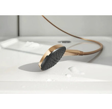 Handdouche in brons-optiek met een zwarte straalplaat, liggend op een witte douchebak met waterdruppels.