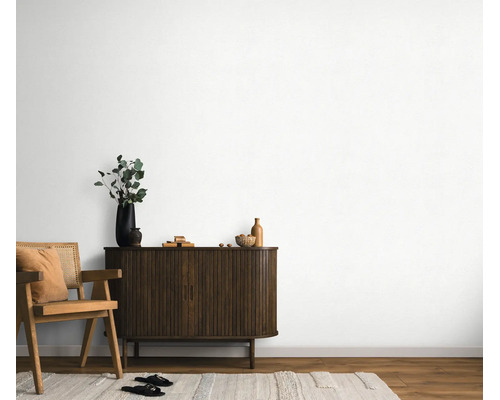 Woonruimte met witte muurafwerking, donkerhouten dressoir met latjesdeuren en een stoel in rotandesign op een licht tapijt.