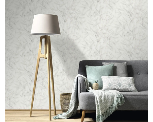 Woonkamer met vliesbehang met bladmotief, grijze bank, houten vloerlamp en woonaccessoires in een gezellig en modern interieur.
